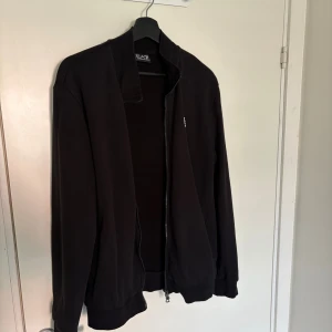 Svart bomberjacka från EA7 Emporio Armani - Svart bomberjacka från EA7 Emporio Armani med dragkedja framtill och ribbade muddar. Jackan har en stilren design med EA7-logga på bröstet och vita ränder på ryggen. Perfekt för dig som gillar en clean och sportig look.