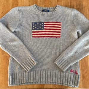 Grå stickad tröja Polo Ralph Lauren - Snygg grå stickad tröja från Polo Ralph Lauren med amerikansk flagga broderad på bröstet och RL-broderi i rött vid nederkanten. Tröjan har rund hals, långa ärmar och breda ribbade muddar. Perfekt för dig som gillar klassisk och cool stil.