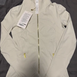 Vit tröja från Lululemon med matchande tights - Snygg vit vindjacka från Lululemon med guldig dragkedja och smickrande sömmar i strl 2. Jackan har lång ärm, hög krage och två fickor med dragkedja. Perfekt för dig som gillar sportig och clean stil. Materialet är mjukt och stretchigt syntet. Tightsen är i storlek 4. 