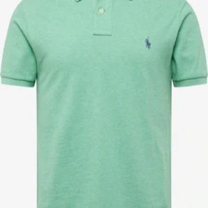 Grön Ralph Lauren Piké  - Fräsch grön piké från Ralph Lauren med klassisk krage och två knappar framtill. Piken har korta ärmar och den ikoniska lila Polo-loggan broderad på bröstet. Lite längre i ryggen! 