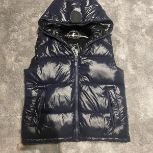 Save The Duck väst med huva mörkblå - Snygg mörkblå dunväst från Save The Duck med glansig finish och huva. Västen har två fickor med dragkedja och coolt tryck på insidan. Perfekt för lager-på-lager och streetstyle. Stängs med dragkedja framtill. Mycket sparsamt använd priset är ej hugget i sten!                                    nypris: 2700 