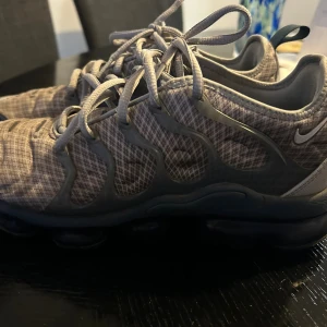 Nike Air Vapormax Plus rutiga sneakers - Nike Air Vapormax Plus sneakers i grått med rutigt mönster och transparenta detaljer på sidorna. Skorna har snörning, en unik bubblig sula och är tillverkade i syntetmaterial. Perfekta för dig som gillar en sportig och modern look. Litet hål på främre sko annars inget annan märkvärdigt 
