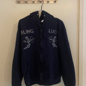 Mörkblå hoodie från Bling Lucid - Mörkblå hoodie med dragkedja och huva. Framsidan har texten 'BLING LUCID' och två cupider i blingade stenar. Mjuk insida, fickor framtill och justerbar dragsko i huvan. Perfekt för dig som gillar streetstyle och detaljer som sticker ut. 