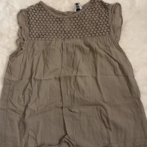 Beige blus med spetsdetalj - Säljer en beige blus från H&M med ärmlös design och volangkant vid axlarna. Ovanpartiet har ett snyggt spetsmönster och knäppning bak i nacken. Blusen är tillverkad i ett lätt och luftigt material, perfekt för varmare dagar.