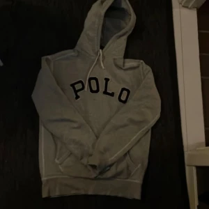 Grå hoodie från Polo Ralph Lauren - Snygg grå hoodie från Polo Ralph Lauren med stor POLO-logga i svart på bröstet. Tröjan har huva med snörning och en klassisk känguruficka framtill. Perfekt för en chill och avslappnad stil.