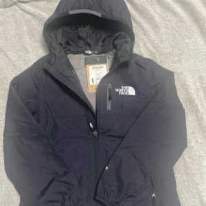 Svart vindjacka från The North Face- storlek S i barn storlek  - Svart vindjacka från The North Face med huva och dragkedja framtill. Jackan har meshfoder, bröstficka med dragkedja och logga på bröstet. Perfekt för blåsiga dagar och enkel att slänga på sig när vädret svänger. i storlek S is barn storlek                                                      frakta samma dag 🚚 
