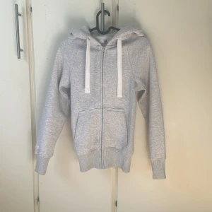 Ljusgrå hoodie från Russedress XXS - Ljusgrå hoodie från Russedress i storlek XXS. Tröjan har dragkedja framtill, två fickor, huva med vita snören och ribbade muddar. Perfekt för chill dagar och har en clean look med diskret logga på ärmen.