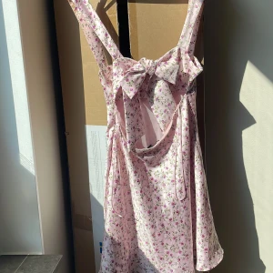 Blommig ljusrosa kortklänning med rosett - Supersöt ljusrosa kortklänning med små blommor i rosa och grönt. Klänningen har breda axelband, öppen rygg med stor rosett och omlott-detalj bak. Materialet är mjukt och lätt, perfekt för varma dagar. 