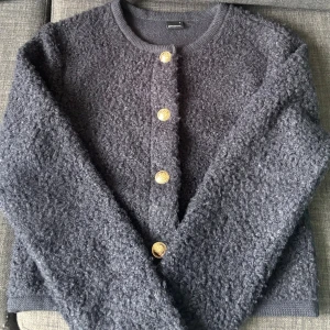 Svart kofta från Gina Tricot med guldknappar - Mysig svart kofta från Gina Tricot med lång ärm och fyra dekorativa guldknappar framtill. Koftan är stickad med en fluffig, strukturerad yta och har en rak, något croppad passform. Perfekt för lager-på-lager under kyliga dagar.