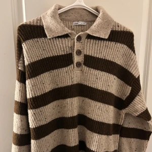 Randig stickad pikétröja från Zara - Säljer en grovstickad pikétröja från Zara med breda bruna och beige ränder. Tröjan har krage, tre knappar framtill och långa ärmar. Perfekt för dig som gillar retrovibbar och vill ha en mysig, oversized look.