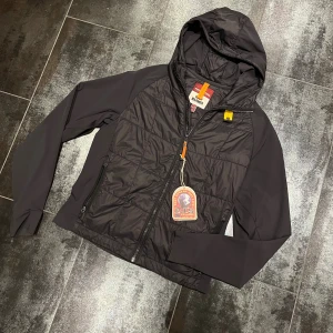 Svart Parajumpers jacka XS med huva - Svart Parajumpers jacka i modellen Shrike med huva och dragkedja. Jackan har quiltad front, släta ärmar och detaljer i orange och gul. Tillverkad i syntetmaterial och passar perfekt för kyliga dagar. Snygg och sportig look med praktiska fickor. Nypris 4199kr