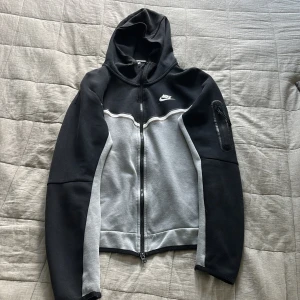 Grå och Svart nike tech - Nike hoodie i svart och grå med hel dragkedja framtill, huva och vit Nike-logga på bröstet. Tröjan har långärmad design, ficka med dragkedja på ena ärmen och sportig look. Perfekt för chill eller träning.