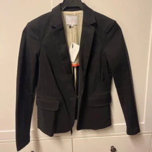 Svart kavaj från Coster Copenhagen - Stilren svart kavaj från Coster Copenhagen i klassisk modell med smal krage och två fickor framtill. Kavajen har en snygg, figurnära passform och är tillverkad i ett mjukt material som ger en skön känsla. Perfekt för dig som gillar minimalistisk och modern stil. Kavajen är aldrig använd och har prislapp kvar. 