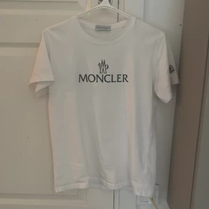 Vit Moncler t-shirt med logga - Vit t-shirt från Moncler med svart logotyp tryckt på bröstet och Moncler-märke på ärmen. Klassisk rund hals och korta ärmar. Tillverkad i mjuk bomull för skön passform. Perfekt för dig som gillar stilrena designer och exklusiva märken. T-shirten är i storlek M men är liten i storlek vilket gör att den sitter som S. Priset är absolut inte spikat. Bara att höra av sig om det finns något intresse!