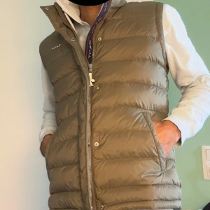  GANT Väst mycket bra skick - Snygg olivgrön dunväst från GANT med quiltad design och hög krage. Västen har dragkedja och tryckknappar framtill, samt två praktiska fickor. Insidan har ett mörkt foder och GANT-logga på bröstet och insidan av öppningen. Perfekt för lager-på-lager under kyliga dagar. Den är knappt buren och då är i mycket gott skick. Den kan bäras som S men passar lite bättre för M