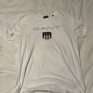 Vit GANT t-shirt med logga. Aldrig använd - Vit t-shirt från GANT med klassisk rund hals och tryckt logga på bröstet. På framsidan finns ett stort GANT-tryck och ett emblem med stjärnor och ränder i blått och rött. Skön passform och tillverkad i mjuk bomull.