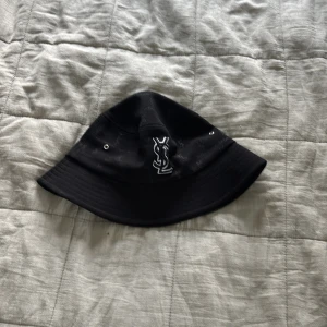 Svart YSL bucket hat  - Snygg svart bucket hat från YSL med broderad vit logga framtill och små YSL-mönster över hela hatten. Klassisk form med metallöglor på sidorna för ventilation. Perfekt för dig som vill ha en stilren och trendig accessoar.