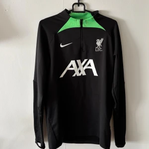 Liverpool FC långärmad träningströja Nike 23/24 - Liverpool FC träningsjacka 23/24 från Nike med lång ärm, svart bas och gröna detaljer vid halsen. Stor vit AXA-logga på bröstet och broderat LFC-märke. Materialet är mjukt och stretchigt, perfekt för träning eller match. Halv dragkedja framtill för enkel ventilation. Tröjan har även hål vid tummarna så att man kan värma händerna. Perfekt för hösten och våren. Tröjan är storlek S men passar även M. Finns det frågor och funderingar är det bara att höra av sig/team F&S.