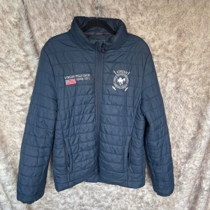 Mörkblå quiltad jacka från Vinson Polo Club - Säljer en mörkblå quiltad jacka från Vinson Polo Club med broderade detaljer och amerikansk flagga på bröstet. Jackan har dragkedja framtill, ståkrage och  Perfekt för kyliga dagar