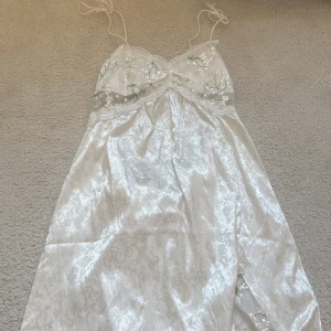 Vit blommig satin klänning med broderi - Supersöt vit klänning i satin med tunna axelband och blommigt mönster. Överdelen har broderade detaljer med små vita blommor och gröna blad, samt spetskant. Kjolen har ett subtilt blommigt mönster i tyget som ger en lyxig känsla. Perfekt för sommarens alla tillfällen!