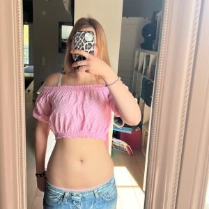 Rosa offshoulder croppad topp - Säljer en söt rosa offshoulder topp med puffiga korta ärmar och smockdetaljer vid både halsringning och nederkant. Toppen är croppad och har en lätt strukturerad bomullskänsla med små broderade prickar. Perfekt för varma dagar och en trendig look.