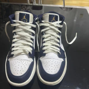 Nike Air Jordan 1 Mid Navy/Vit - Säljer ett par Nike Air Jordan 1 Mid i marinblått och vitt skinn med klassisk Jordan-logga i guld på plösen och sidan. Skorna har snörning, perforerad tå och den ikoniska swoosh-loggan. Perfekta för dig som vill ha en stilren och sportig look.