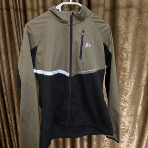 Svart och olivgrön vindjacka Newline - Sportig vindjacka från Newline i svart och olivgrönt med hel dragkedja, bröstficka med dragkedja och reflekterande detalj över bröst och rygg. Jackan har hög krage och är tillverkad i lätt syntetmaterial, perfekt för utomhusaktiviteter.