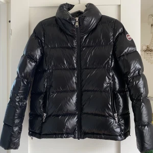 Colmar jacka  - Säljer en svart, glansig pufferjacka från Colmar med hög krage och dragkedja framtill. Jackan har två sidofickor med dragkedja, quiltad design och snörning nedtill. Perfekt för kalla dagar och har en snygg, modern look.