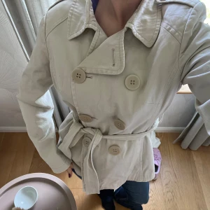 Beige trenchcoat med bälte och knappar - Snygg beige trenchcoat med klassisk krage, dubbelknäppning och matchande knappar. Storlek ”14”, skulle säga att det är en kappa i storlek XS/S! 