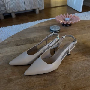 Beige pumps med låg klack från Duffy - Snygga beige pumps från Duffy med spetsig tå och låg klack. Skorna har öppen häl med justerbart spänne och är tillverkade i syntetmaterial. Perfekta för dig som gillar stilrena och eleganta skor med en modern touch. Pumpsen är endast använda en gång så skicket är som nya.