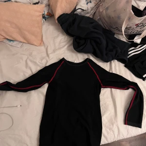 Svart sporttröja från H&M med röda detaljer - Svart långärmad sporttröja från H&M med röda linjer längs ärmar och axlar. Tröjan har en enkel, tight passform och är tillverkad i ett mjukt syntetmaterial som passar perfekt för träning eller chill dagar.