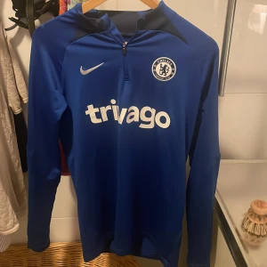 Chelsea långärmad tröja! - Chelsea FC långärmad träningströja från Nike i blått med klubbmärke och Trivago-tryck på bröstet. Slim fit-modell i Dri-FIT-material som andas, halv dragkedja framtill och sportig look. Perfekt för dig som älskar fotboll och vill ha en snygg fotbollströja! Köpt direkt från UNISPORT, fullt äkta!