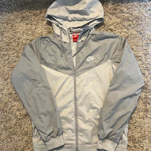 Grå Vit Nike vindjacka med huva herr  - Snygg grå vindjacka från Nike med vit logga på bröstet. Jackan har huva med snörning, hel dragkedja och meshfoder på insidan. Mycket bra skick, inga märkvärdiga defekter. Postas antigen samma dag som köp eller dagen därpå🙌💯