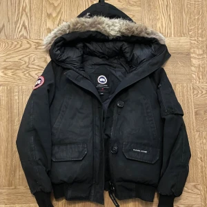 Svart dunjacka från Canada Goose - Säljer en svart dunjacka från Canada Goose med huva och snygg fuskpäls runt kanten. Jackan har flera fickor, ribbade muddar och dragkedja med knappslå. Klassisk patch på ärmen och varm vaddering, perfekt för kalla dagar.