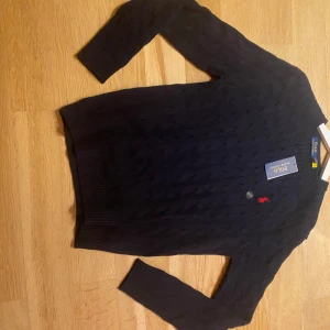 Mörkblå kabelstickad tröja Polo Ralph Lauren - Mörkblå kabelstickad tröja från Polo Ralph Lauren med klassisk röd broderad logga på bröstet. Tröjan har rund halsringning, ribbade muddar och är långärmad. Perfekt för lager-på-lager och snygg till jeans eller kjol.