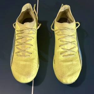 Gula Puma Ultra Ultimate AG/FG fotbollsskor - Säljer ett par gula Puma Ultra fotbollsskor med snörning och svart logga på sidan. Skorna har en smal och sportig form, mönstrad ovandel och dobbar i genomskinlig plast för bästa grepp på planen. Lätta och flexibla, perfekta för snabba rörelser.