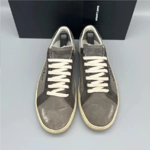 Saint Laurent grå sneakers i canvas - Grå sneakers från Saint Laurent i canvas med råa kanter och vit sula. Skorna har vita snören och svart Saint Laurent-tryck på sidan. Snygg vintagekänsla och klassisk låg modell som passar till allt. Perfekt för dig som gillar streetstyle och exklusiva märken.