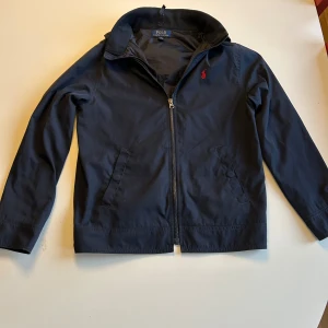 Mörkblå vindjacka från Polo Ralph Lauren - L barnstolen men sitter som s Inga tydliga defekter. Snygg mörkblå vindjacka från Polo Ralph Lauren med klassisk krage, dragkedja framtill och två snedställda fickor. Jackan har det ikoniska röda Polo-logot broderat på bröstet och är perfekt för dig som vill ha en stilren och tidlös look.