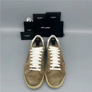 Saint Laurent sneakers i mocka - Snygga Saint Laurent sneakers i beige mocka med vit häl och vita snören. Klassisk låg modell med handskriven logga på sidan. Perfekta för dig som gillar stilrena och exklusiva skor med en twist.