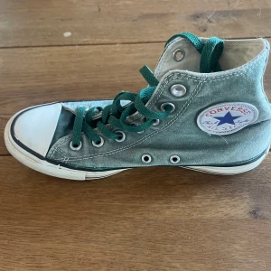 Gröna Converse All Star high-tops - Klassiska Converse All Star high-tops i grön canvas med vita gummidetaljer och gröna snören. Skorna har rund tå, platt sula och den ikoniska loggan på sidan. Perfekta för dig som vill ha en tidlös och cool sneakerstil.