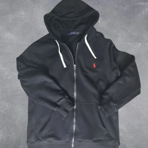 Polo Ralph Lauren zip hoodie - Svart zip hoodie från Polo Ralph Lauren med dragkedja, vit snörning i huvan och röd broderad logga på bröstet. Mjuk bomullskvalitet och känguruficka framtill. Perfekt för en chill och stilren look.