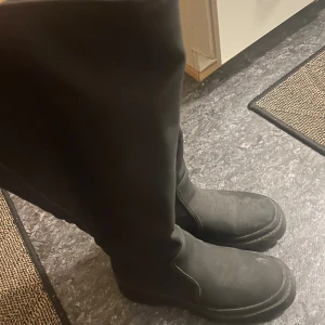 Svarta höga boots i mocka-look - Säljer ett par svarta höga boots med platt sula och rund tå. Skorna har en stilren design och är tillverkade i material som liknar mocka. Perfekta för dig som vill ha en enkel men snygg look till höst och vinter.