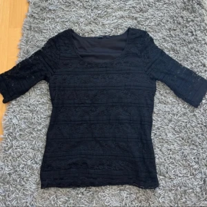 Svart spetsblus från Lindex - Svart blus från Lindex i storlek S med halvtransparent spets och rund halsringning. Blusen har trekvartsärm och ett fodrat liv, vilket ger en snygg kontrast mellan spetsmönstret och det täckta tyget. Perfekt för dig som gillar detaljer och stilren look.