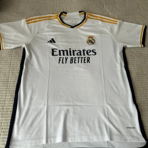 Real Madrid vit fotbollströja Adidas XL - Vit Real Madrid fotbollströja från Adidas i storlek XL. Tröjan har svarta och gula detaljer på krage och axlar, samt klubbmärke och sponsortryck på bröstet. Tillverkad i lätt och ventilerande polyester med korta ärmar och slim fit-passform.