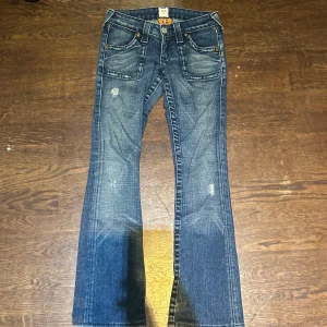 True Religion 504 bootcut jeans blå - Snygga blå True Religion 504 jeans med bootcut och slitna detaljer. Klassisk femficksmodell med kontrastsömmar och markerade bakfickor. Jeansen har låg midja och är tillverkade i kraftigt denim med cool tvättad look.