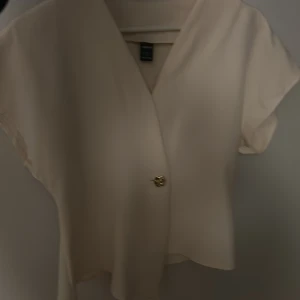Beige omlottblus från SHEIN - Säljer en stilren beige omlottblus från SHEIN i storlek S. Blusen har korta ärmar, v-ringning och en snygg guldig knapp framtill. Perfekt för dig som gillar en clean och enkel look med en twist. Materialet känns lätt och flowy.