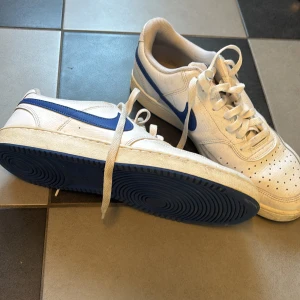 Nike Court Vision vita/blå sneakers - Nike Court Vision sneakers i vitt skinn med blå swoosh och detaljer. Klassisk låg modell med snörning, perforerad tå och platt sula. Bekväm mesh runt ankeln och insida. Kan förhandla om priset 