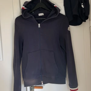 Mörkblå hoodie från Moncler - Snygg mörkblå hoodie från Moncler med dragkedja framtill, huva med röd, vit och blå rand samt broderad logga på ärmen. Tröjan har känguruficka och ribbade muddar med Monclers signaturränder. Perfekt för dig som gillar stilrena streetwear-plagg.
