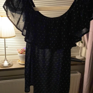 Mörkblå blus med volang  - Supersöt mörkblå blus med små vita mönster och bred volang runt axlarna. Tröjan är luftig och har offshoulder-stil, perfekt för varma dagar. Tunt, lätt material som ger en flowy känsla. Men man kan styla den på sitt sätt vid axlarna.