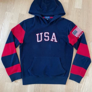 Mörkblå hoodie från Polo Ralph Lauren - Mörkblå hoodie från Polo Ralph Lauren med röd-vita USA-detaljer och broderad amerikansk flagga på ärmen. Tröjan har huva, känguruficka och breda röda ränder på ärmarna. Tillverkad i mjuk fleece som är perfekt för kyliga dagar.